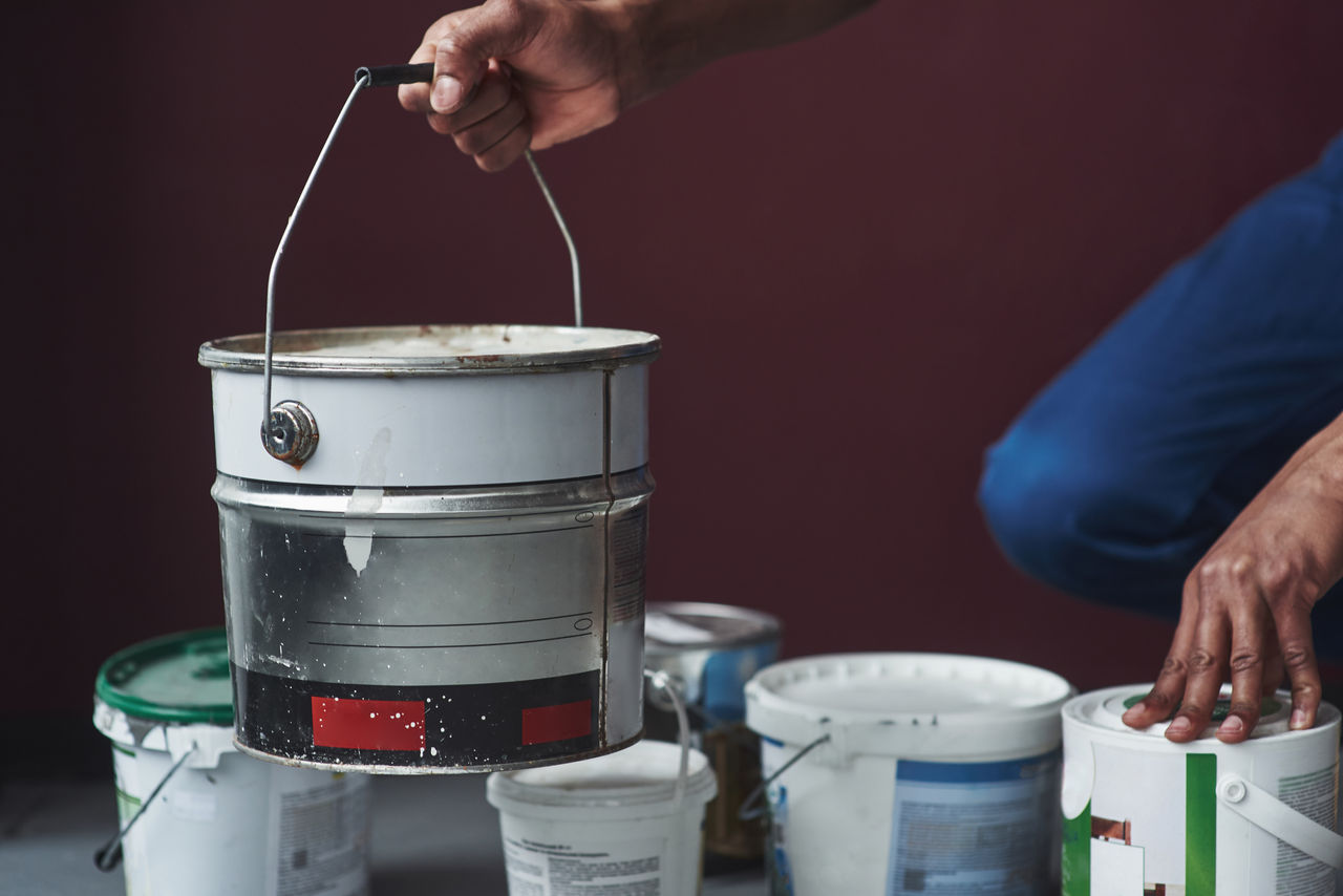 A hand holding a bucket of paint with several other buckets resting on the floor